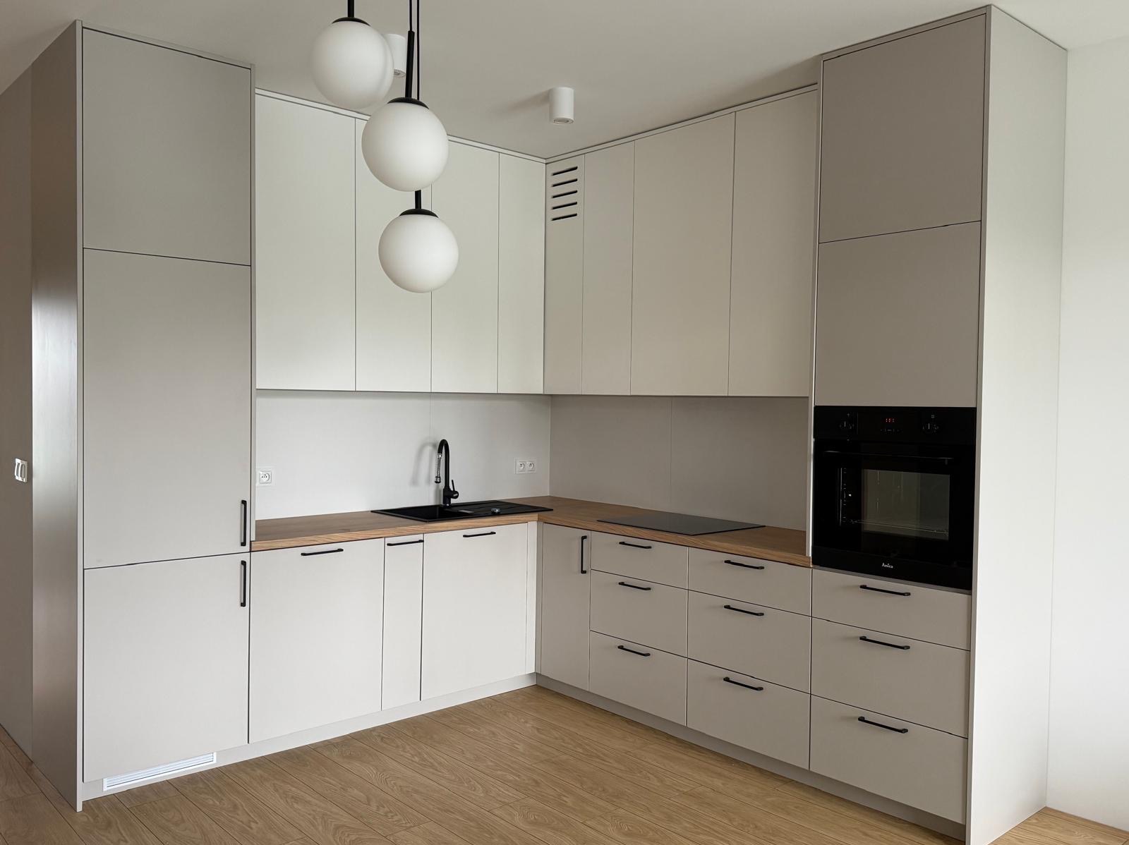 Jasna kuchnia z frontami bez uchwytów, drewnianym blatem, czarnym zlewem i baterią oraz trzema okrągłymi lampami wiszącymi.
