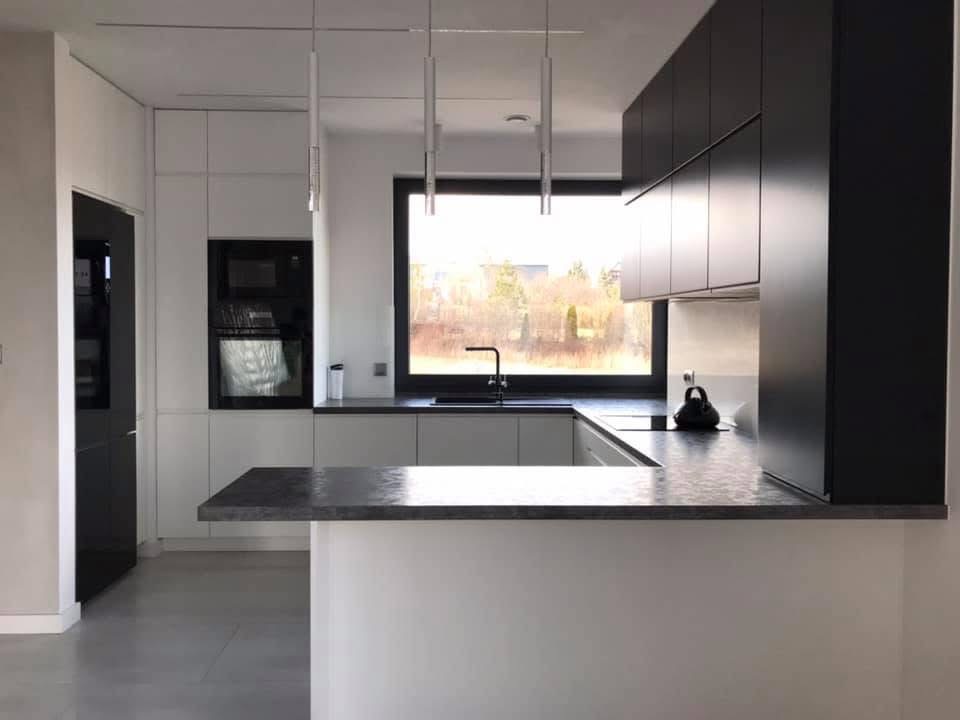Nowoczesna kuchnia z czarno-białymi szafkami, wyspą z szarym blatem i minimalistycznym oświetleniem. Widok na zewnątrz przez okno.