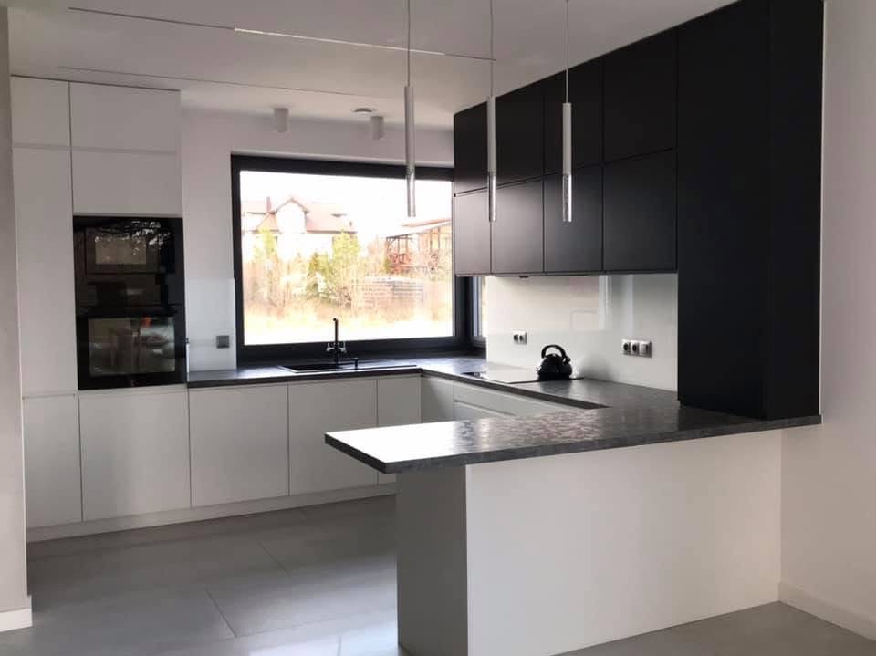 Nowoczesna kuchnia w bieli i czerni z wyspą, oświetleniem punktowym i dużym oknem z widokiem na zieleń. Minimalistyczny design z czajnikiem na blacie.