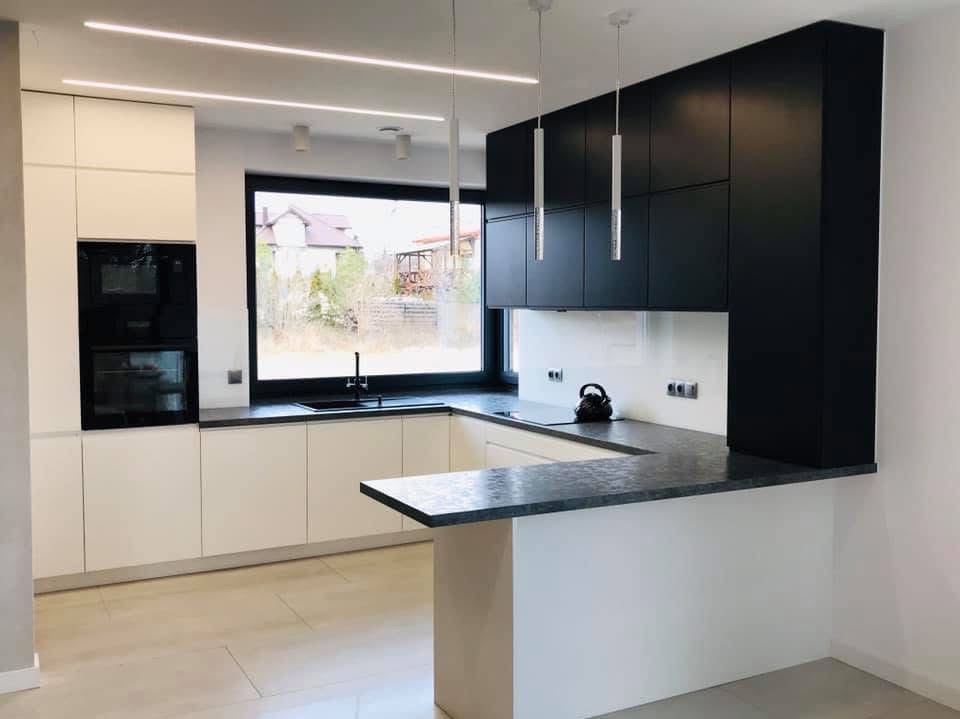 Nowoczesna kuchnia w bieli i czerni z wyspą, oświetleniem LED i dużym oknem z widokiem na ogród. Minimalistyczny design z elementami dekoracyjnymi.