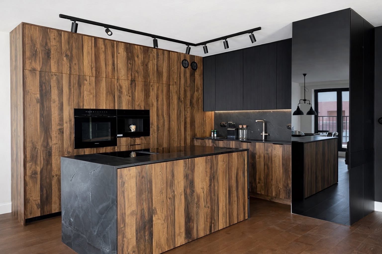 Nowoczesna kuchnia z wyspą: fronty z ciemnego drewna o wyraźnym usłojeniu, czarne szafki górne i blat z ciemnego kamienia. Oświetlenie punktowe na szynoprzewodzie.