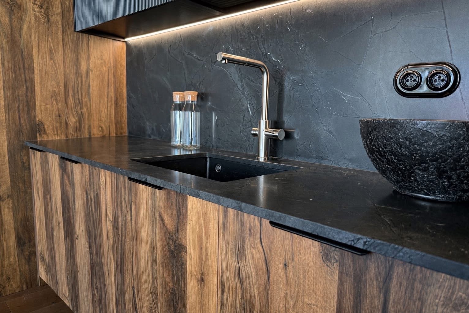 Nowoczesna kuchnia z drewnianymi frontami, czarnym blatem i marmurową ścianą. Bateria chromowana, czarna misa i szklane butelki dodają elegancji.