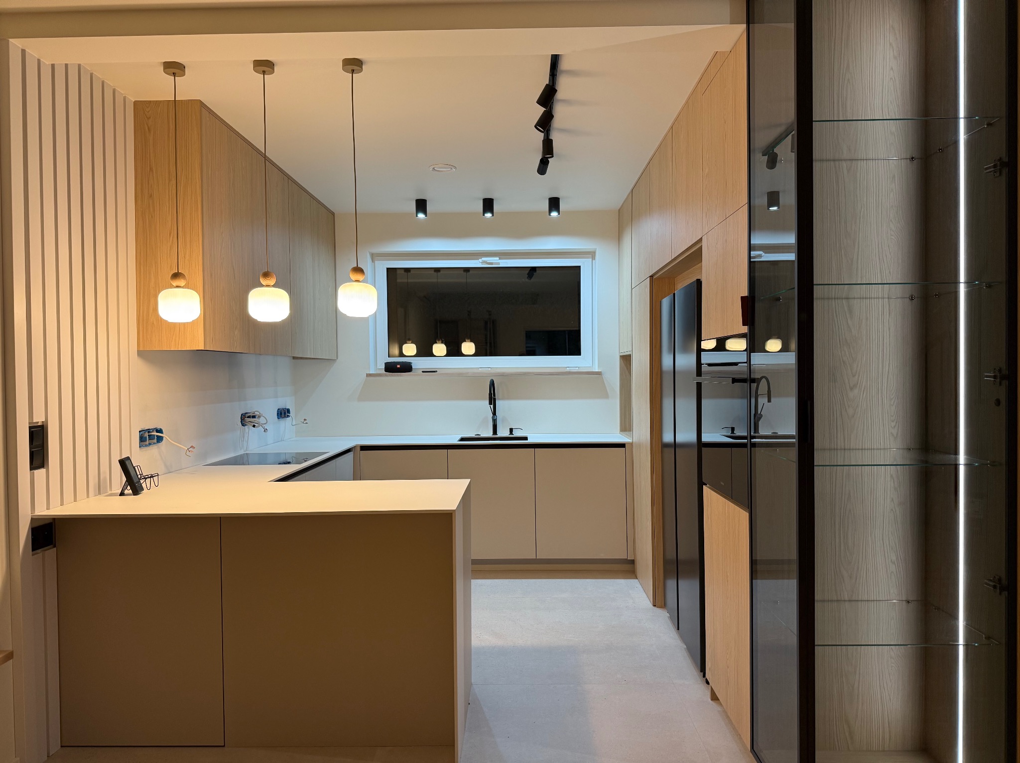 Nowoczesna kuchnia z drewnianymi frontami, wyspą i oświetleniem punktowym. Minimalistyczny design, jasne kolory, szklane witryny. Widoczne gniazdka elektryczne.