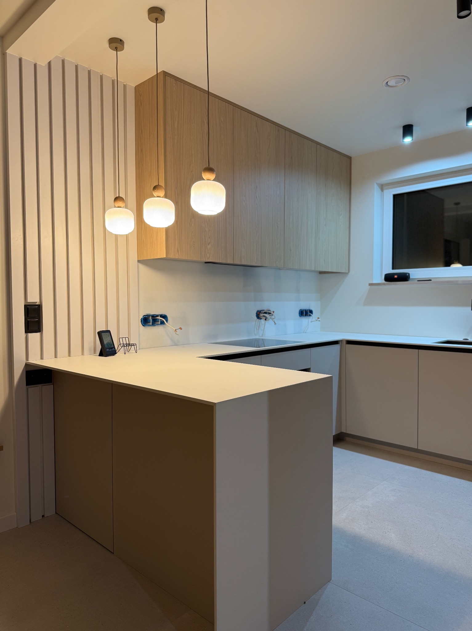 Nowoczesna kuchnia z drewnianymi szafkami wiszącymi i wyspą z blatem. Na blacie czytnik e-booków i miniaturowe krzesło. Ściana z pionowymi panelami dekoracyjnymi.