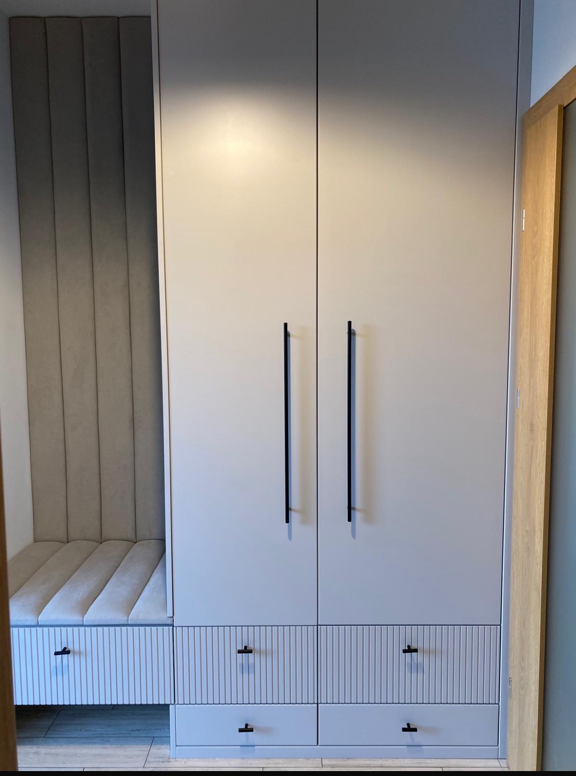 Jasnoszara szafa z siedziskiem i pionowymi frezami na frontach szuflad, długie czarne uchwyty, tapicerowane oparcie siedziska, widok z dołu.