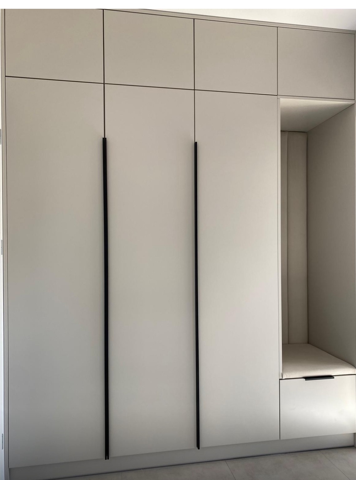 Jasnoszara szafa wnękowa z czarnymi, długimi uchwytami i wbudowanym siedziskiem z tapicerowanym oparciem, oświetlonym od góry.