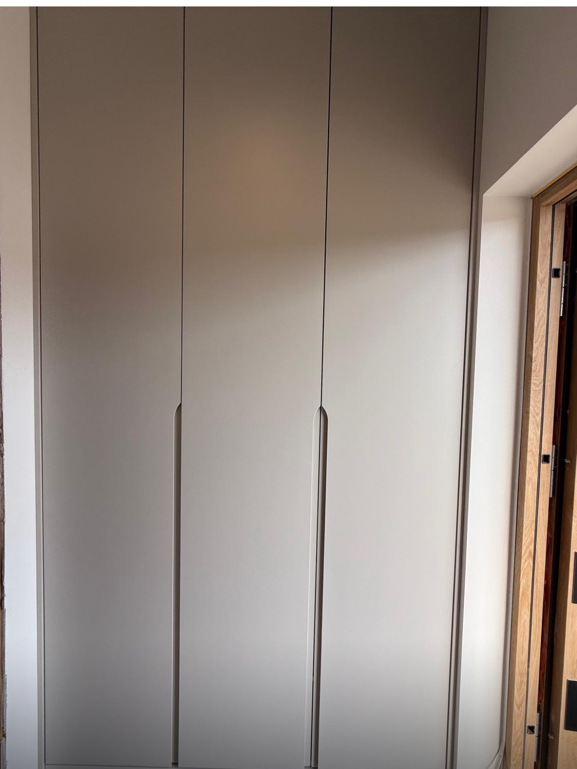 Minimalistyczna, biała szafa wnękowa z trzema drzwiami, zintegrowanymi uchwytami i subtelnym oświetleniem, wkomponowana w jasne wnętrze.