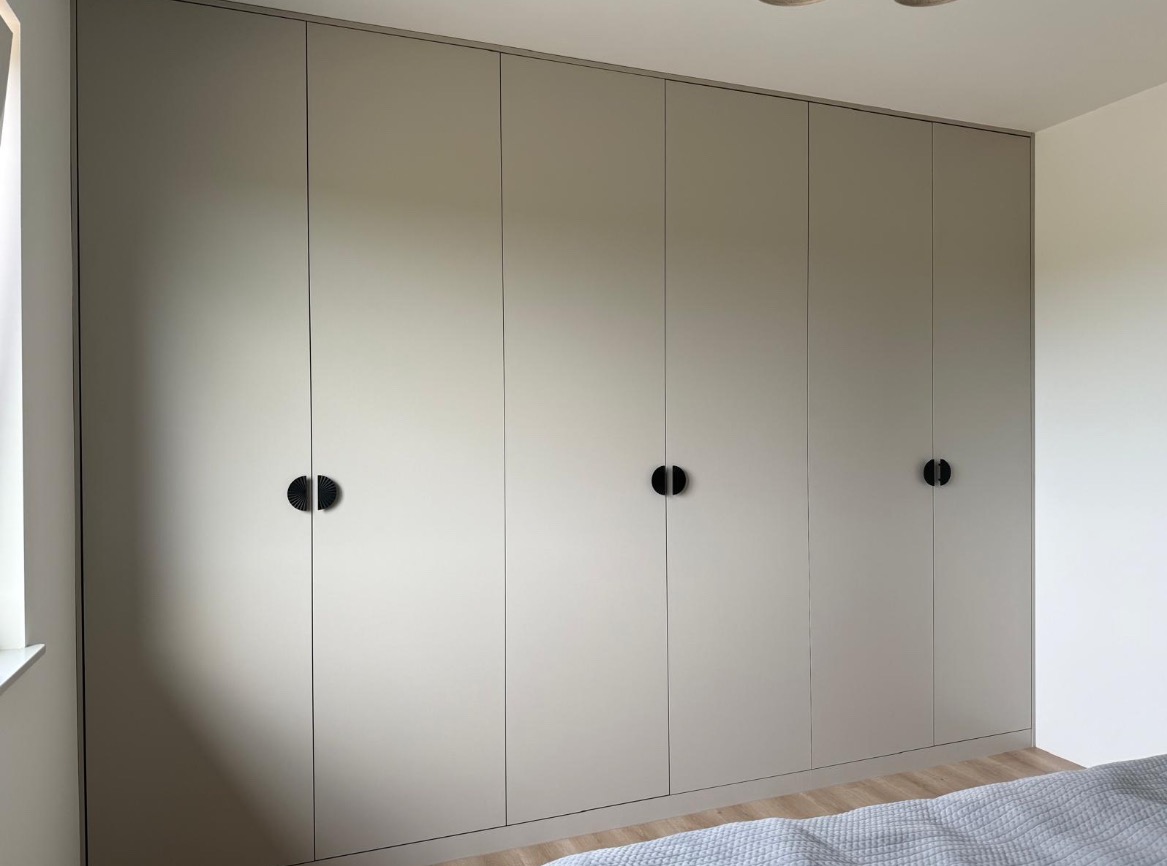 Beżowa szafa wnękowa z minimalistycznymi, okrągłymi uchwytami na tle jasnej ściany i podłogi. Widoczne detale wykończenia i subtelne oświetlenie.