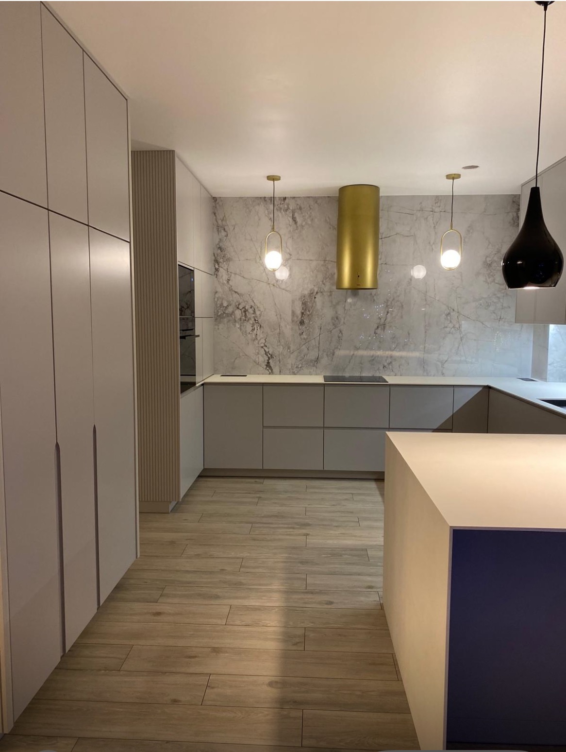 Nowoczesna kuchnia z szarymi frontami, złotym okapem i marmurową ścianą. Drewnopodobna podłoga i minimalistyczne lampy wiszące dopełniają elegancki wystrój.
