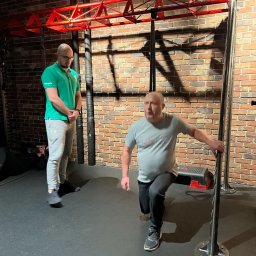 MJP Michał Podolski - Mężczyzna wykonuje przysiad wykroczny, trzymając się metalowej konstrukcji, pod nadzorem trenera w zielonej koszulce polo, na tle ceglanej ściany i czerwonej konstrukcji do ćwiczeń.