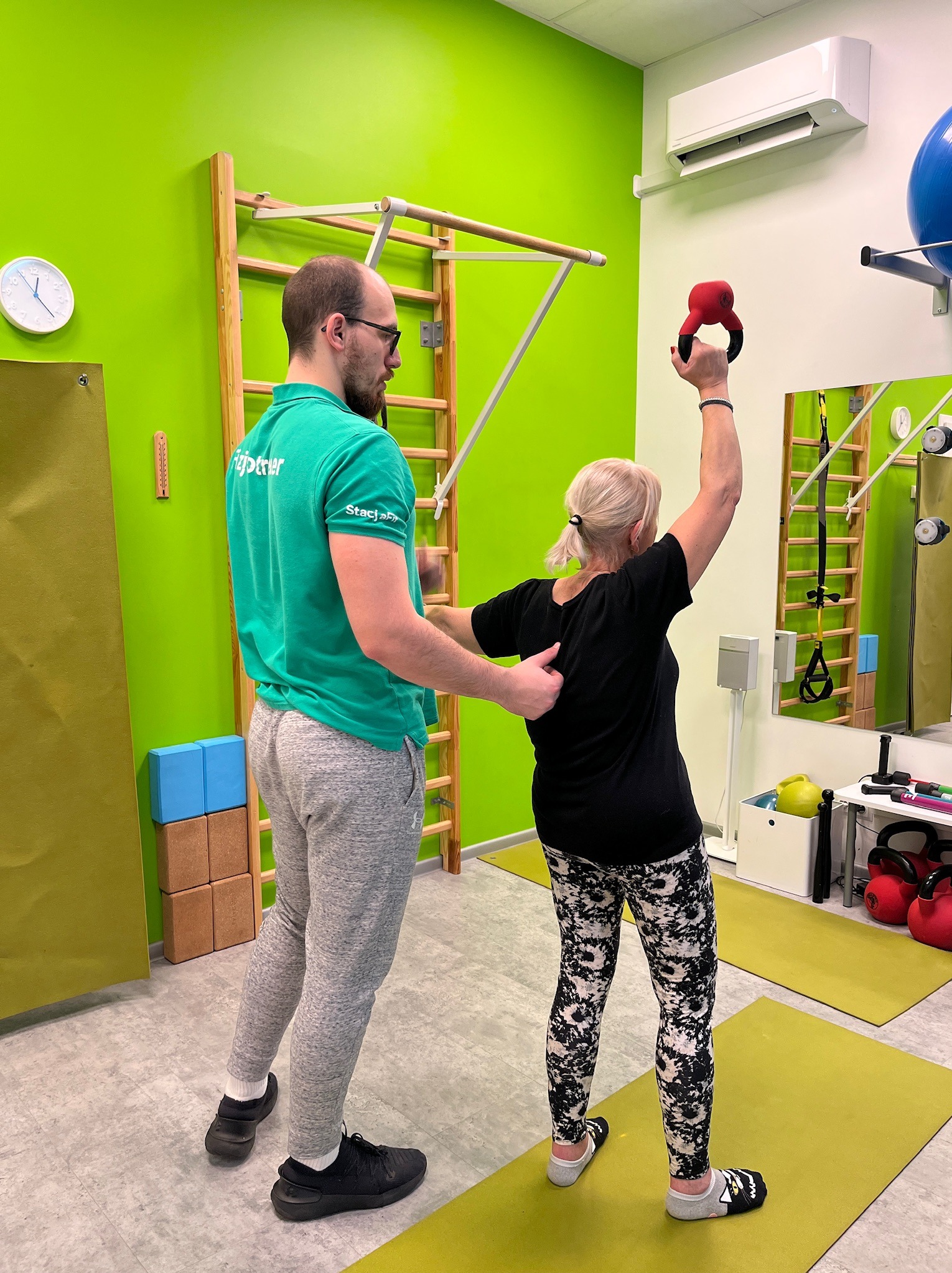 Kobieta ćwiczy z czerwonym kettlebell pod okiem mężczyzny w zielonej koszulce w jasnym pomieszczeniu z drabinką gimnastyczną i lustrem.