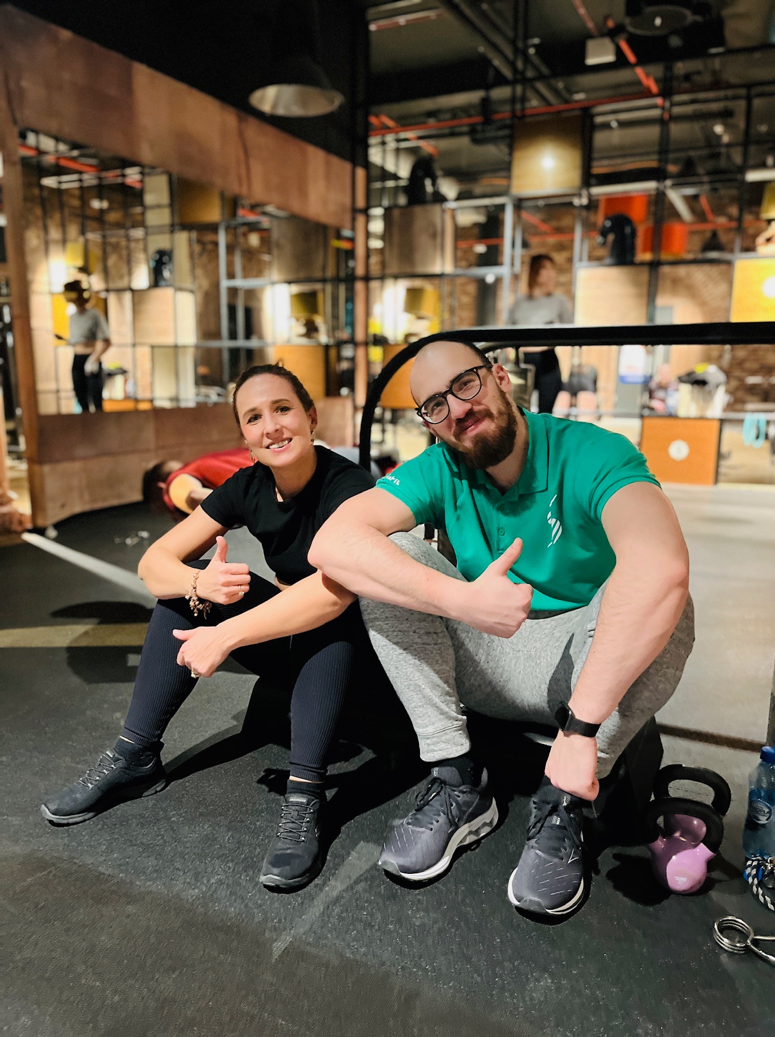 Uśmiechnięta kobieta i mężczyzna w okularach, oboje siedzą na podłodze w siłowni i pokazują kciuki w górę, obok nich leżą kettlebells i butelka wody.