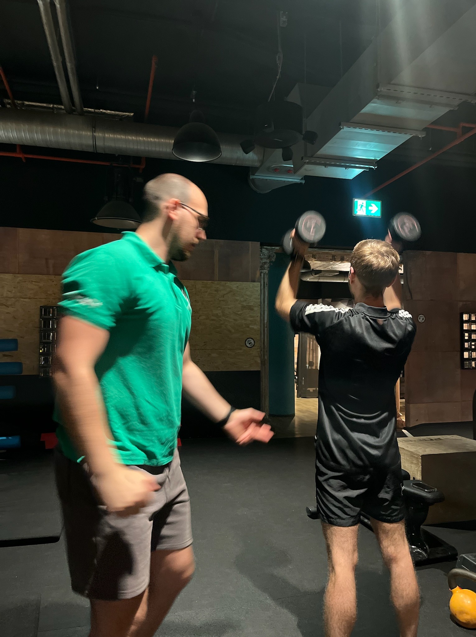 Mężczyzna w zielonej koszulce nadzoruje ćwiczenie unoszenia hantli przez mężczyznę w czarnej koszulce w siłowni z widocznymi rurami wentylacyjnymi pod sufitem.