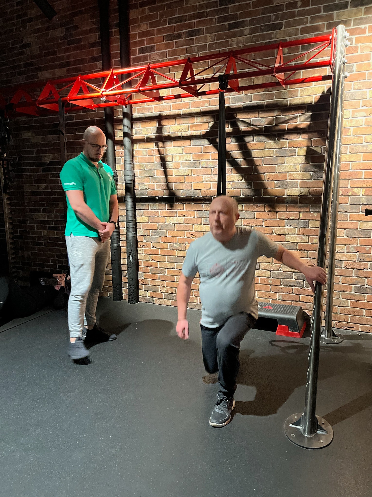 Mężczyzna wykonuje przysiad wykroczny, trzymając się metalowej konstrukcji, pod nadzorem trenera w zielonej koszulce polo, na tle ceglanej ściany i czerwonej konstrukcji do ćwiczeń.