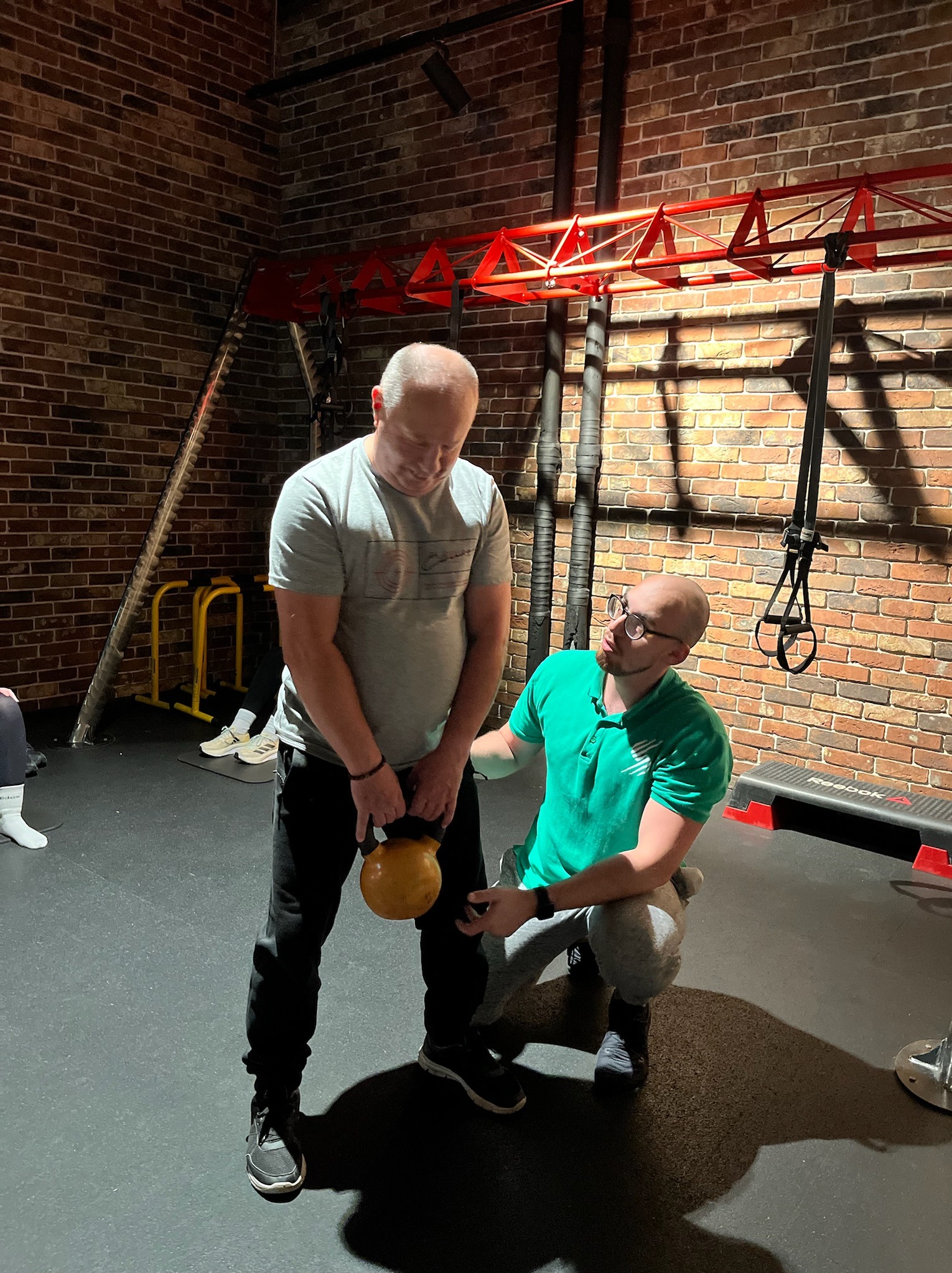 Mężczyzna ćwiczy z odważnikiem kettlebell pod nadzorem trenera w zielonej koszulce, ceglana ściana i sprzęt do ćwiczeń w tle.