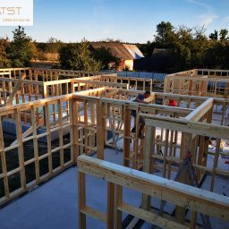 ATST Konstrukcje Drewniane - Wznoszenie drewnianej konstrukcji domu na betonowej płycie fundamentowej, widoczni dwaj pracownicy w trakcie montażu, w tle zabudowania gospodarcze i drzewa.