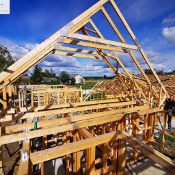 ATST Konstrukcje Drewniane - Budowa domu w technologii szkieletowej: widok na konstrukcję drewnianą z pracownikiem siedzącym na belce w prawym dolnym rogu, błękitne niebo w tle.