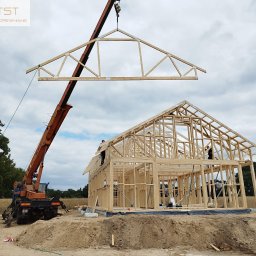 ATST Konstrukcje Drewniane - Montaż drewnianej więźby dachowej na budowie domu szkieletowego przy użyciu dźwigu, widoczni pracownicy na konstrukcji.