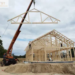 Montaż drewnianej więźby dachowej na budowie domu szkieletowego przy użyciu dźwigu, widoczni pracownicy na konstrukcji.