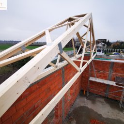 ATST Konstrukcje Drewniane - Konstrukcja drewniana więźby dachowej z metalowymi łącznikami, widok z góry na murowane ściany budynku w trakcie budowy, rusztowanie w tle.