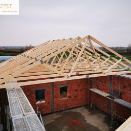 ATST Konstrukcje Drewniane - Wznoszenie drewnianej więźby dachowej na murowanym budynku w trakcie budowy, widoczne rusztowania i elementy mocujące.