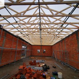 ATST Konstrukcje Drewniane - Wnętrze budynku w trakcie budowy z widoczną konstrukcją więźby dachowej, murowane ściany z czerwonej cegły, rusztowania i narzędzia budowlane na ziemi.