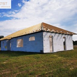 ATST Konstrukcje Drewniane - Surowy stan domu z białych bloczków z widoczną drewnianą konstrukcją dachu, na tle zielonej trawy i drzew.