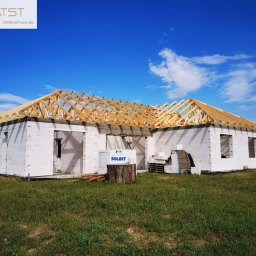 ATST Konstrukcje Drewniane - Surowy stan domu z białych bloczków z drewnianą konstrukcją dachu, na tle błękitnego nieba z chmurami i zielonego trawnika. Widoczne materiały budowlane i pień drzewa.