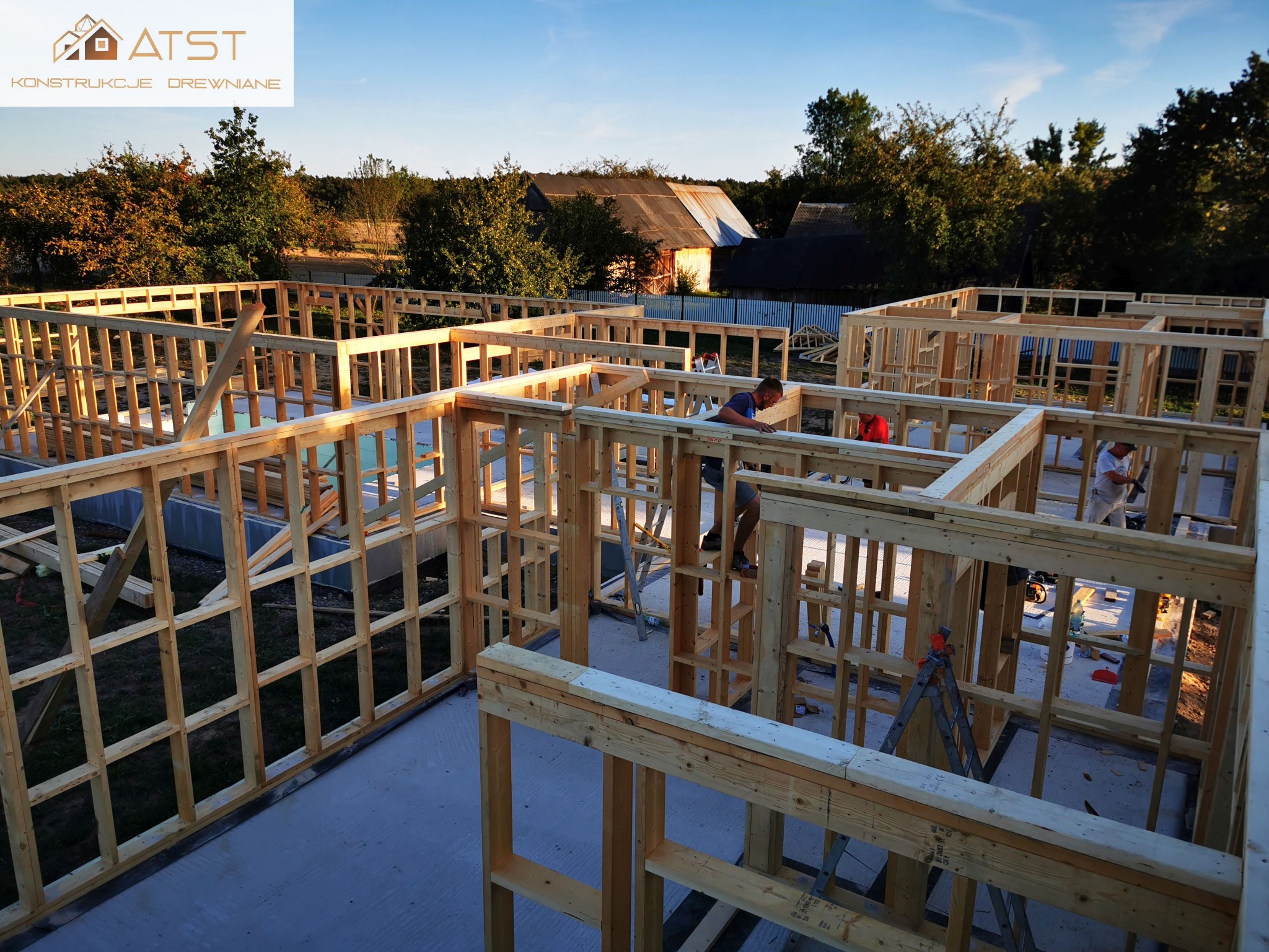 Wznoszenie drewnianej konstrukcji domu na betonowej płycie fundamentowej, widoczni dwaj pracownicy w trakcie montażu, w tle zabudowania gospodarcze i drzewa.