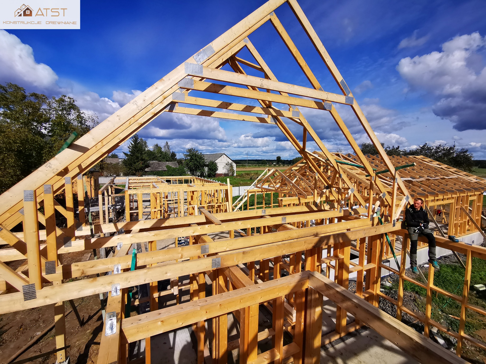 Budowa domu w technologii szkieletowej: widok na konstrukcję drewnianą z pracownikiem siedzącym na belce w prawym dolnym rogu, błękitne niebo w tle.