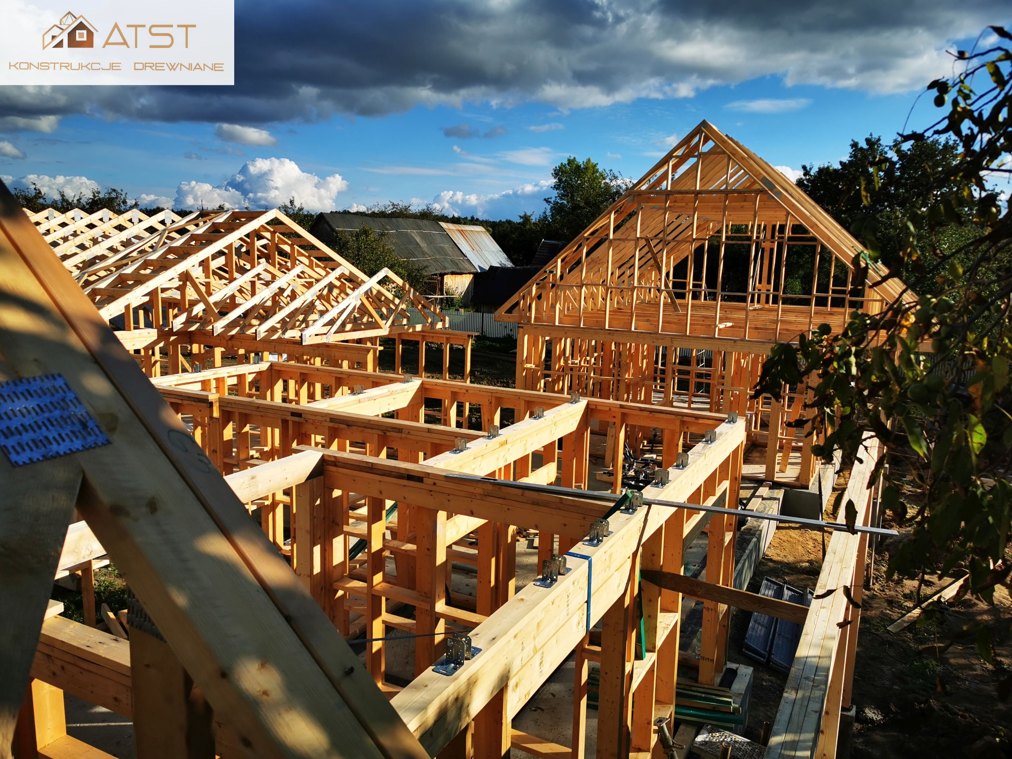 Konstrukcja drewniana domu w trakcie budowy, widoczne elementy szkieletowe, złącza ciesielskie i fragmenty więźby dachowej na tle błękitnego nieba z chmurami.