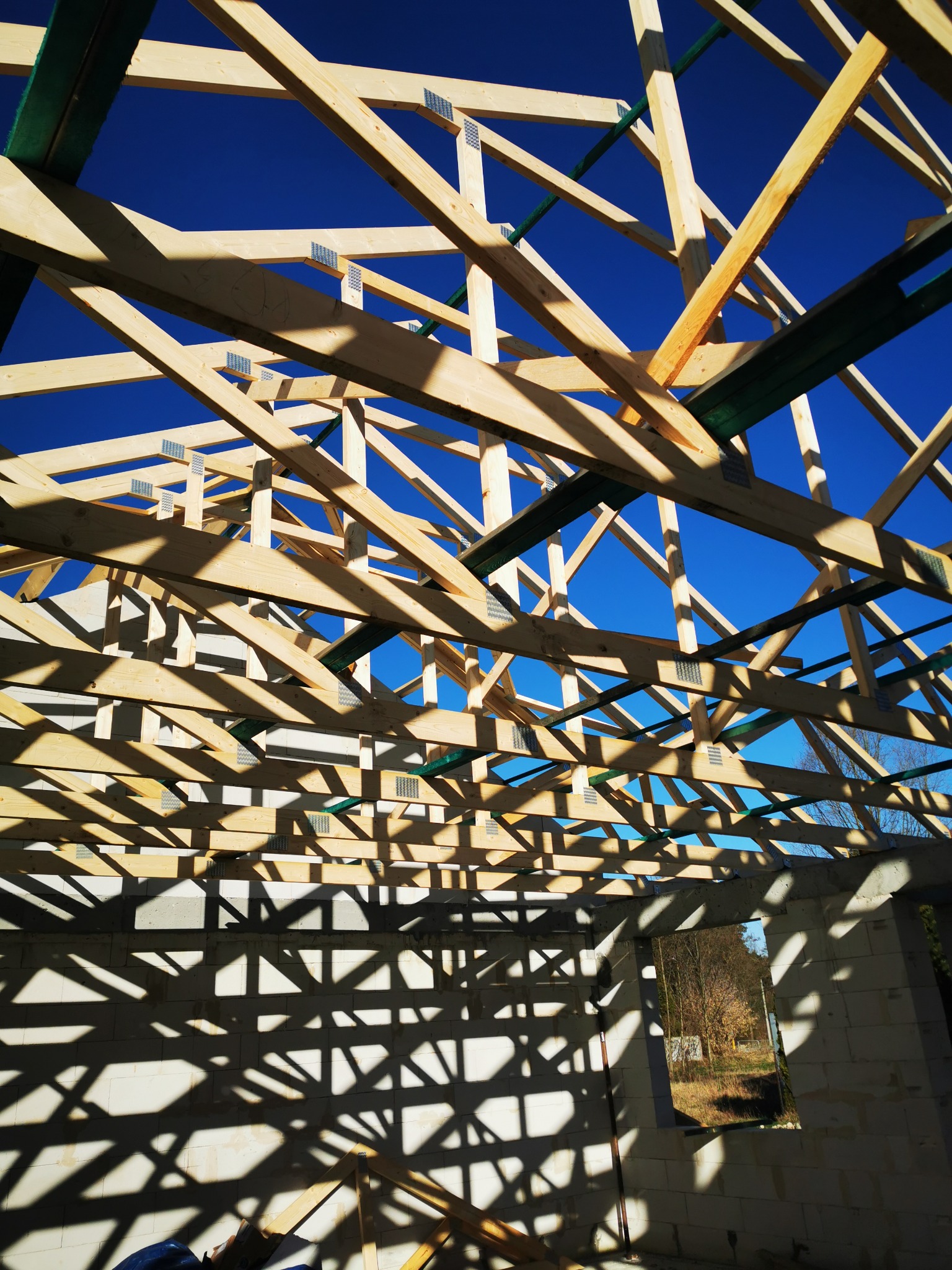 Konstrukcja drewnianej więźby dachowej z widocznymi metalowymi łącznikami, na tle intensywnie niebieskiego nieba, zarys ścian budynku w cieniu.