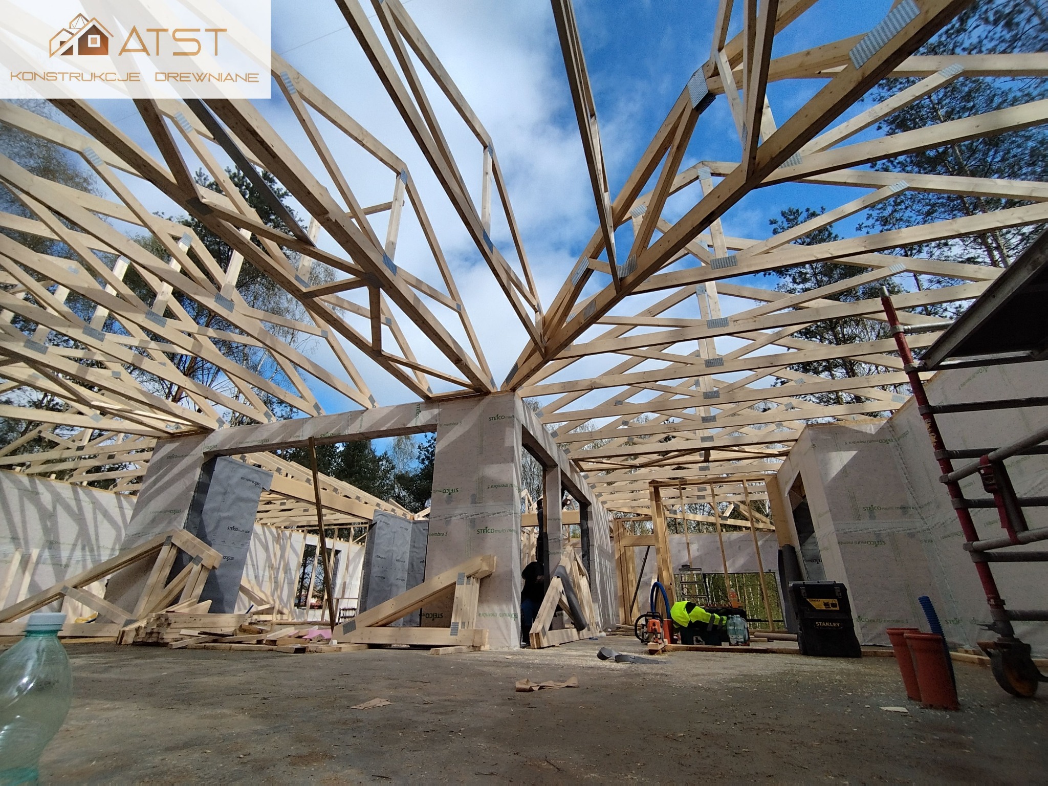 Wnętrze w trakcie budowy domu z widoczną drewnianą konstrukcją dachu, ściany wykończone płytami izolacyjnymi, narzędzia i materiały budowlane na podłodze, widok z poziomu podłogi.