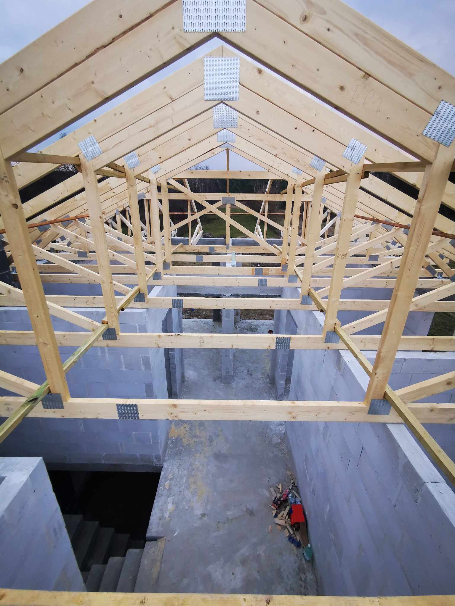 Wnętrze budowanego domu z widoczną konstrukcją więźby dachowej z drewna łączoną metalowymi płytkami, ściany z bloczków betonowych, widok z góry na piwnicę i porozrzucane narzędzia.