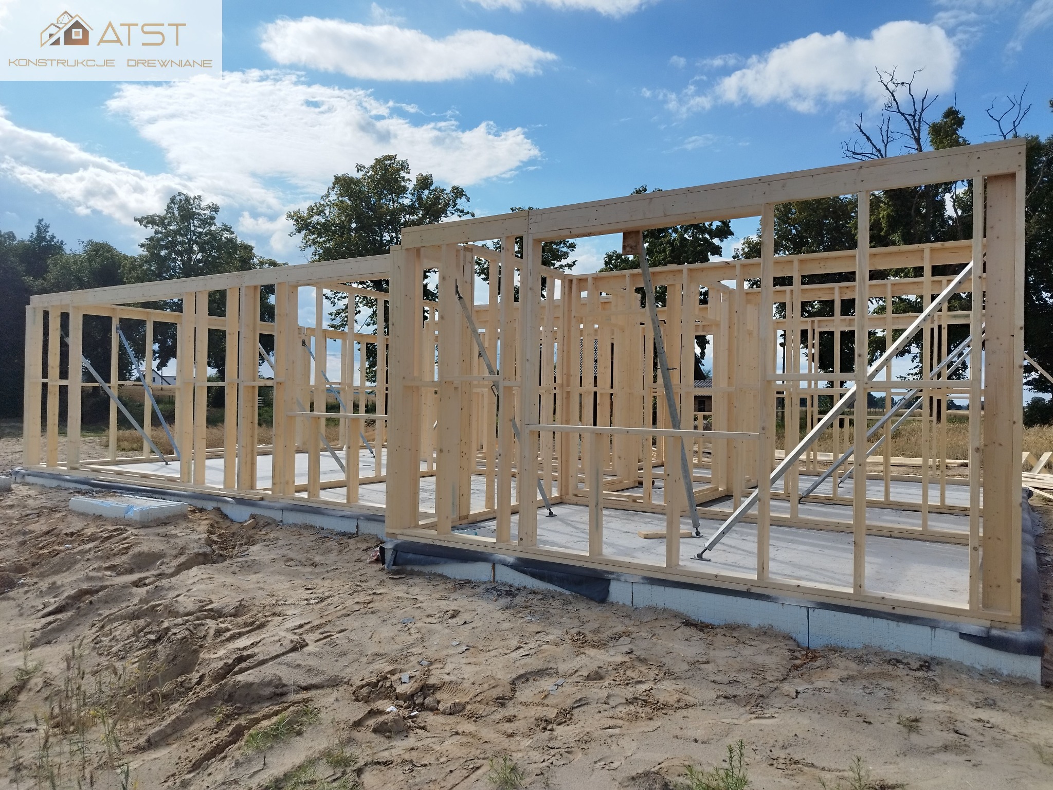 Konstrukcja domu w trakcie budowy, widoczne drewniane ramy ścian na fundamencie, z metalowymi podporami, tło stanowi niebo z chmurami i drzewa.