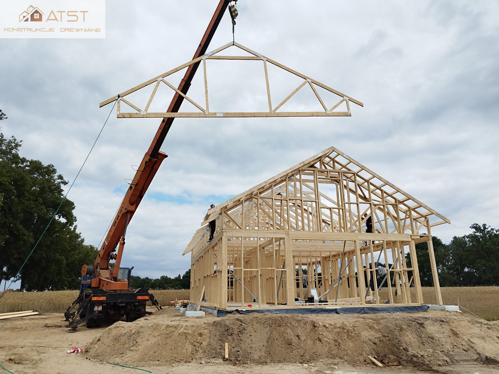 Montaż drewnianej więźby dachowej na budowie domu szkieletowego przy użyciu dźwigu, widoczni pracownicy na konstrukcji.