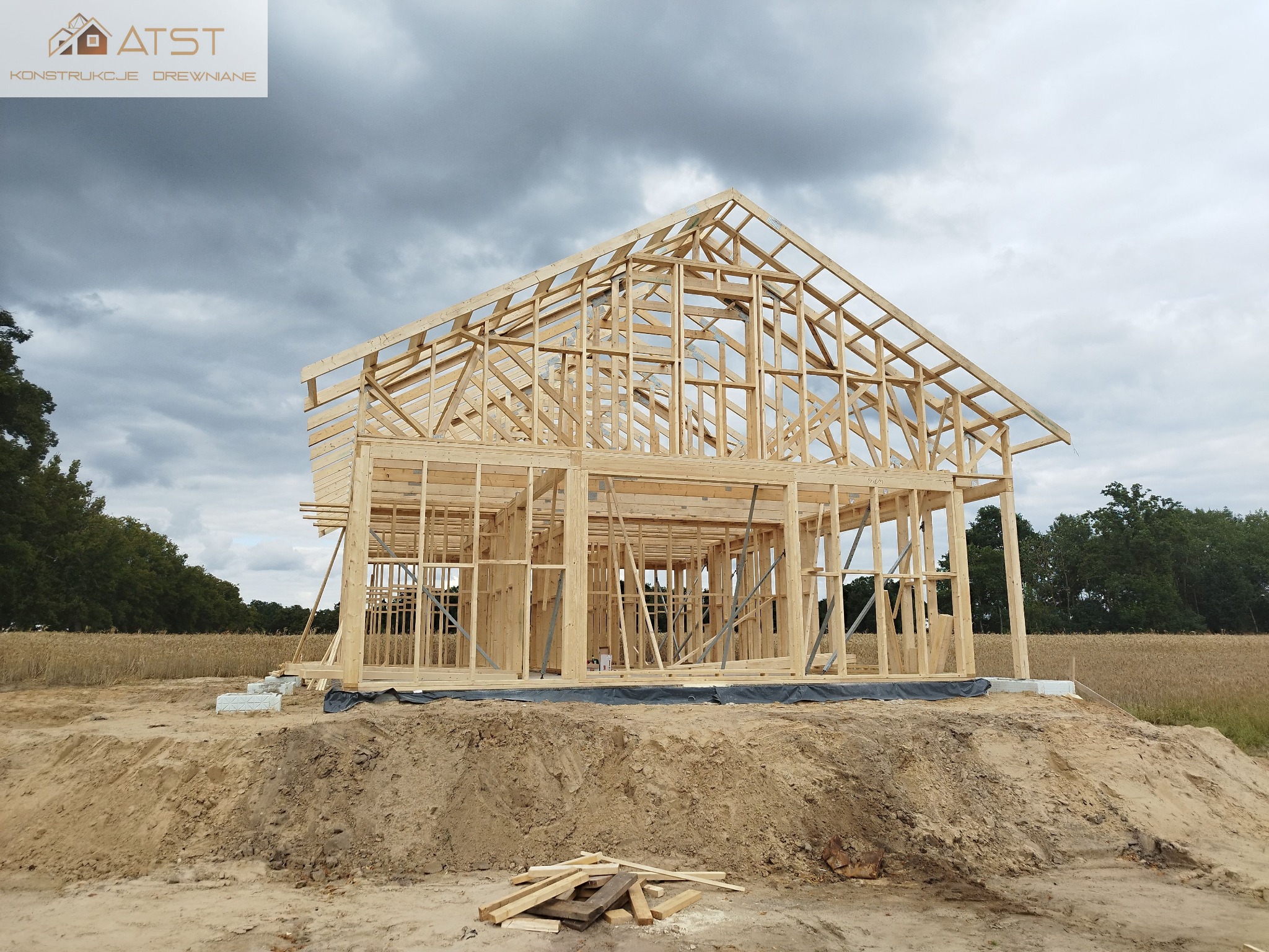 Surowa konstrukcja domu z drewna w trakcie budowy, widoczne elementy szkieletu, podłoże z ziemi, w tle pole i pochmurne niebo.