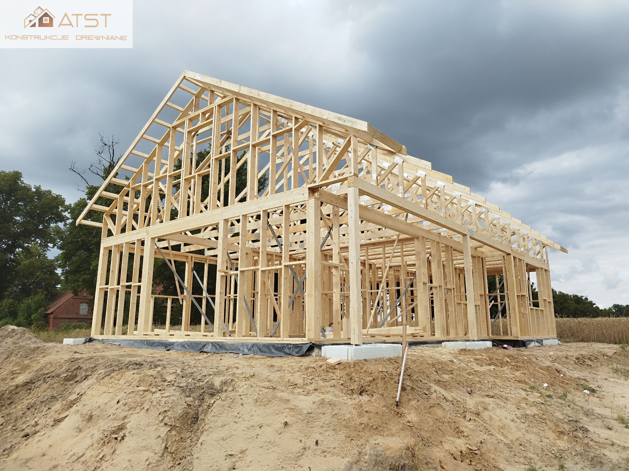 Wznoszenie drewnianej konstrukcji domu na fundamencie, widoczne wiązary dachowe i słupy ścian, na tle zachmurzonego nieba i zieleni drzew.