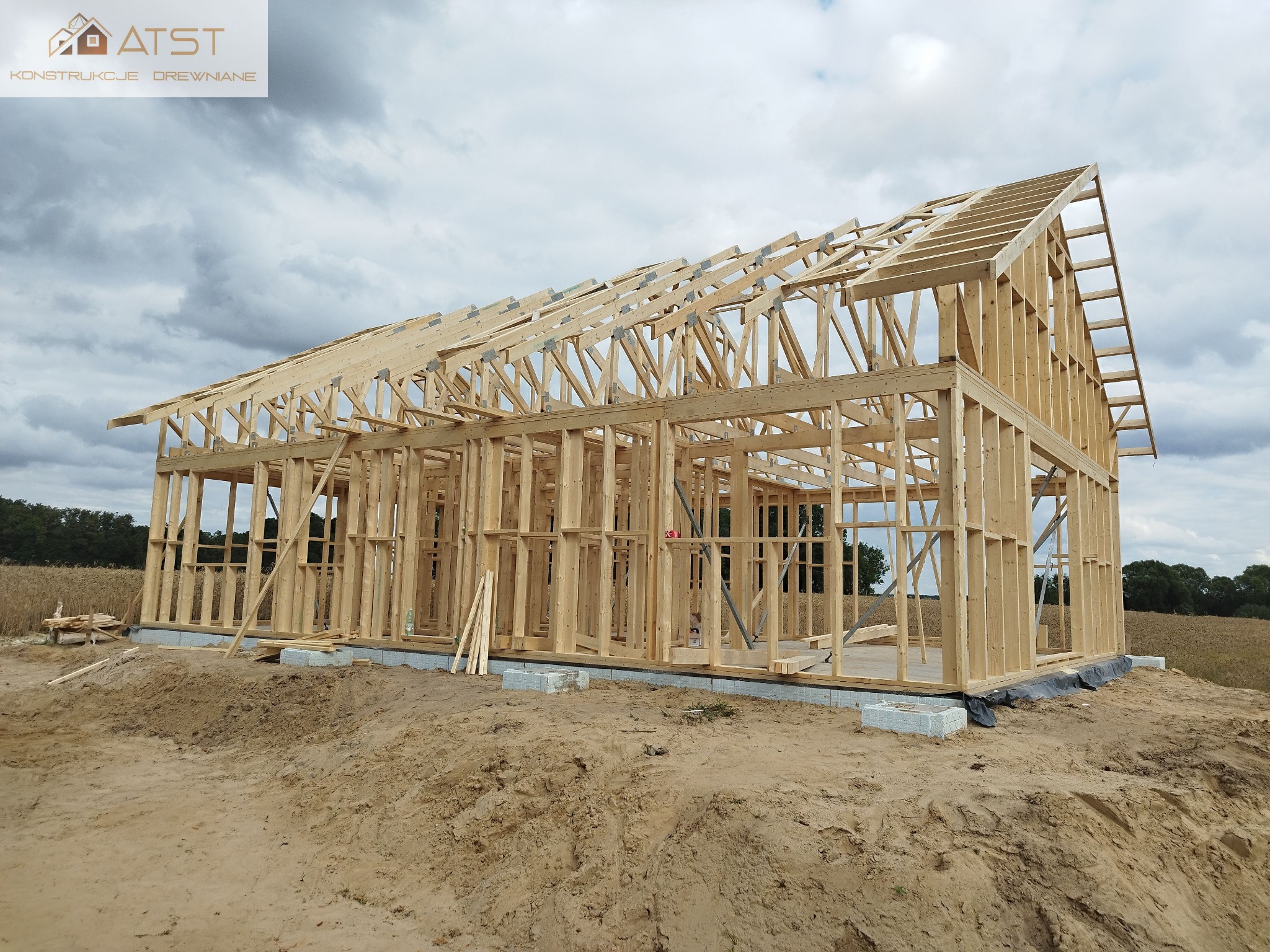Surowy szkielet domu z jasnego drewna na fundamencie z betonowych bloczków, widoczna konstrukcja dachu na tle pochmurnego nieba i pola.