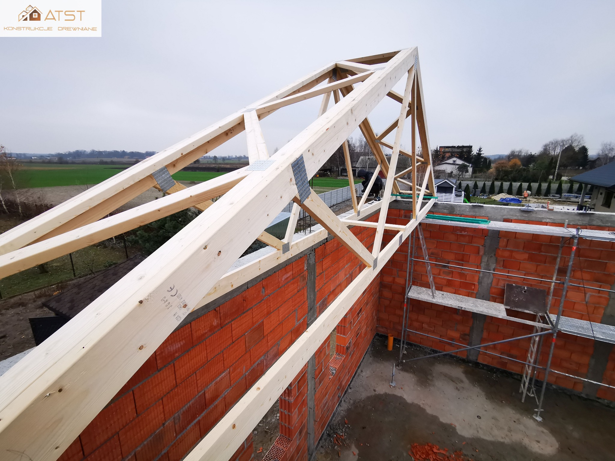 Konstrukcja drewniana więźby dachowej z metalowymi łącznikami, widok z góry na murowane ściany budynku w trakcie budowy, rusztowanie w tle.