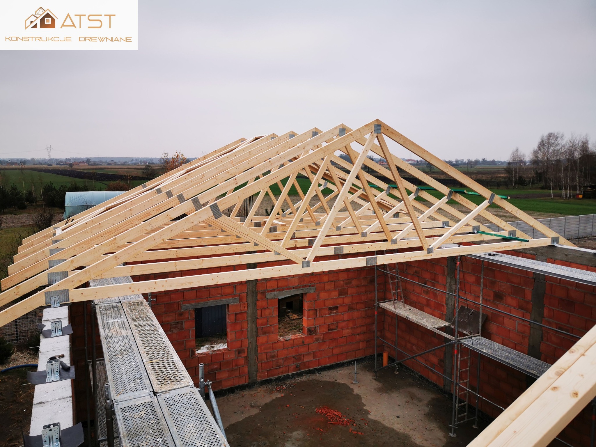 Wznoszenie drewnianej więźby dachowej na murowanym budynku w trakcie budowy, widoczne rusztowania i elementy mocujące.
