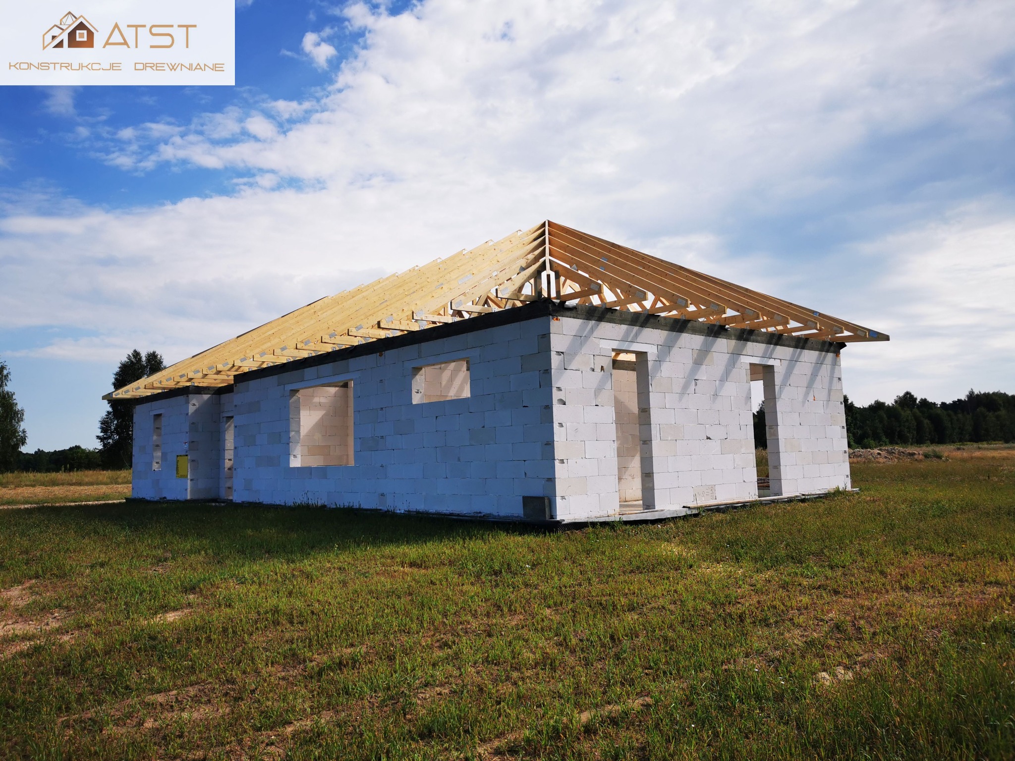 Surowy stan domu z białych bloczków z widoczną drewnianą konstrukcją dachu, na tle zielonej trawy i drzew.