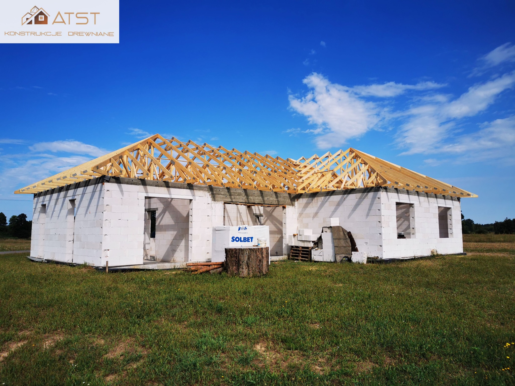 Surowy stan domu z białych bloczków z drewnianą konstrukcją dachu, na tle błękitnego nieba z chmurami i zielonego trawnika. Widoczne materiały budowlane i pień drzewa.