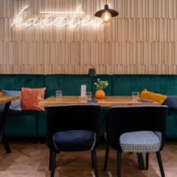 Ustawienie stołu w restauracji: drewniane blaty, krzesła w pepitkę, butelkowa zieleń sofy, dekoracyjna ściana i neon 'Kasztel'. Ciepłe oświetlenie.