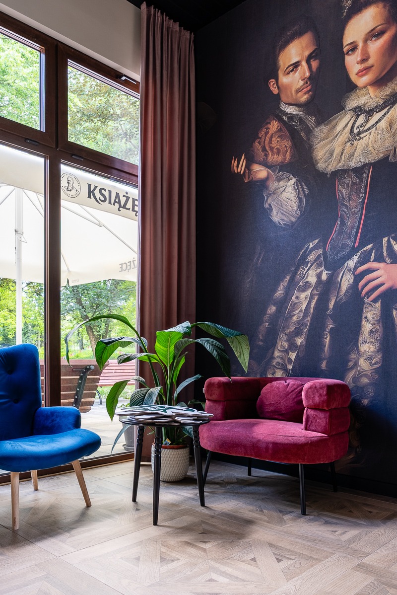 Stylowe wnętrze restauracji: niebieski fotel, bordowa sofa, stolik z menu, okno z widokiem na zewnątrz i mural z parą w strojach historycznych.