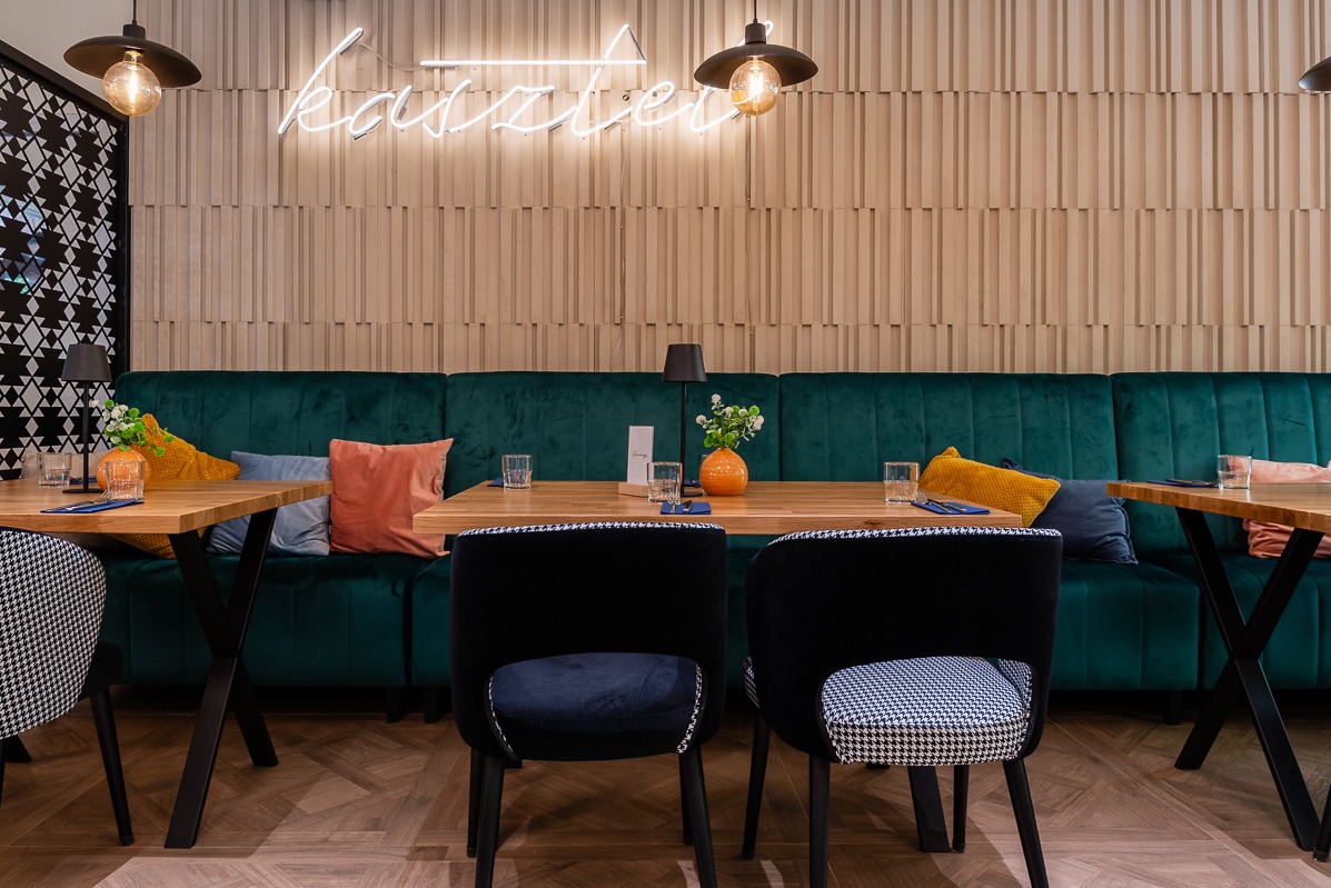 Ustawienie stołu w restauracji: drewniane blaty, krzesła w pepitkę, butelkowa zieleń sofy, dekoracyjna ściana i neon 'Kasztel'. Ciepłe oświetlenie.