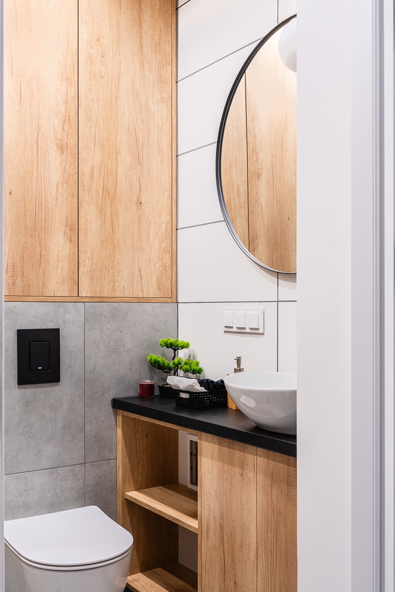 Nowoczesna łazienka z drewnianą szafką i okrągłym lustrem. Umywalka nablatowa, czarny blat, dekoracje. Minimalistyczny styl, jasne płytki, czarne akcenty.
