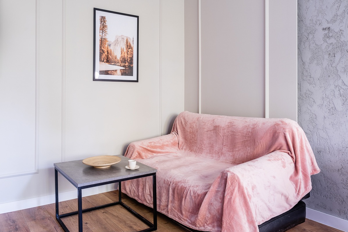 Jasne wnętrze: sofa z różowym pledem, stolik z drewnianą misą i filiżanką, obraz z górskim pejzażem. Dekoracyjna ściana z fakturą betonu.
