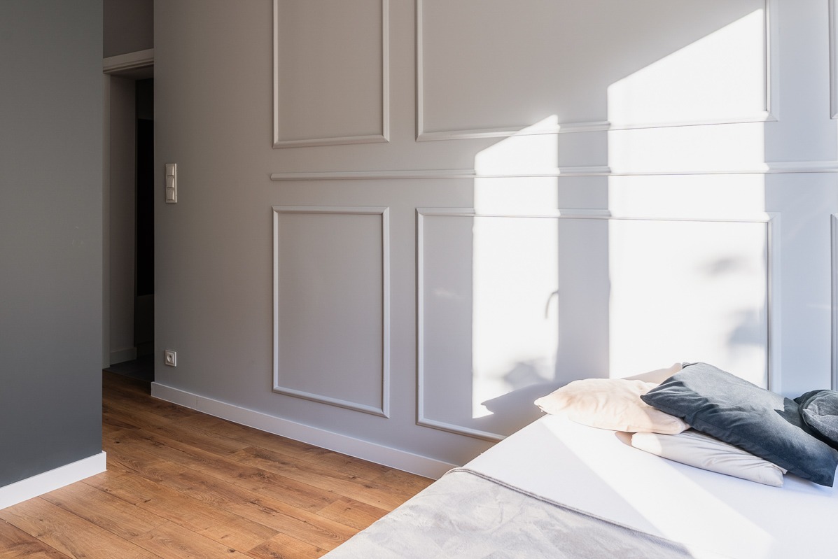 Jasne wnętrze: łóżko z poduszkami w pokoju z panelami ściennymi i drewnianą podłogą. Promienie słoneczne oświetlają ścianę.