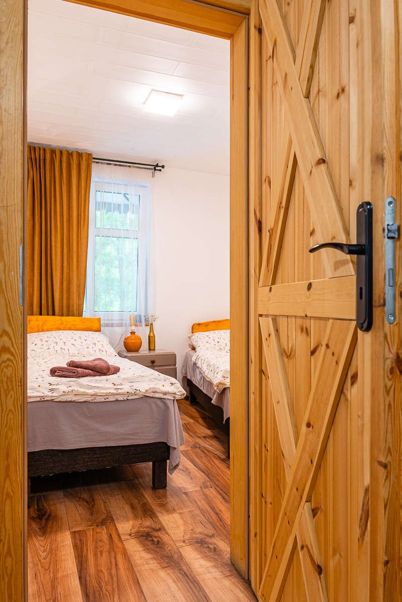 Rustykalny pokój z dwoma łóżkami, widziany przez otwarte drewniane drzwi. Ciepłe kolory, drewniana podłoga, dekoracje na szafce nocnej. Przytulne wnętrze.